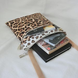 May include: A leopard print crossbody bag with a beige strap. The bag is open, revealing a smartphone, books, and a notebook. The zipper has a white and brown spotted pattern and a tassel.