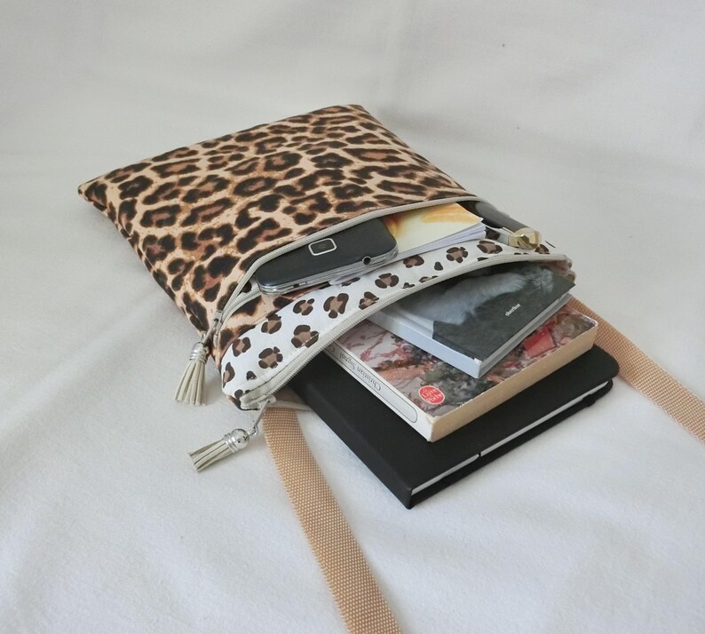 May include: A leopard print crossbody bag with a beige strap. The bag is open, revealing a smartphone, books, and a notebook. The zipper has a white and brown spotted pattern and a tassel.