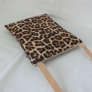 May include: A rectangular, leopard print pouch with a beige strap. The pouch features a zipper closure and a repeating pattern of brown spots on a tan background. The strap is a solid beige color.