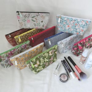 Peut inclure: Une collection de dix trousses rectangulaires en tissu avec fermeture éclair, arborant divers motifs floraux, de papillons et géométriques dans des tons de vert, jaune, rouge, bleu et blanc. Chaque trousse possède un petit pompon sur la tirette.