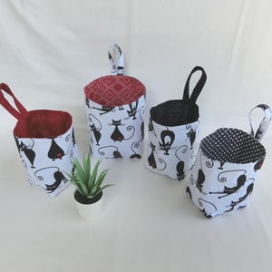 以下が含まれることがあります： 黒い猫のデザインが施された白い布製の吊り下げ収納容器が4つ。容器には、赤、黒、黒と白の市松模様など、さまざまな色の裏地が付いています。