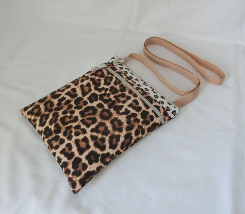 May include: A rectangular crossbody bag with a leopard print pattern in shades of brown and black. The bag has a beige strap and a zipper closure. The bag is made of fabric.