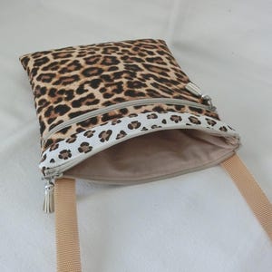 May include: A square crossbody bag with a leopard print pattern in brown and black. The bag has a beige zipper and a matching beige strap. The interior is a solid beige color. The bag is open, revealing the interior.