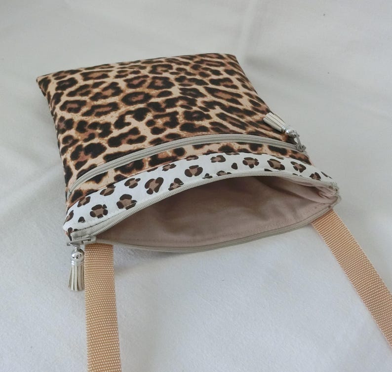 May include: A square crossbody bag with a leopard print pattern in brown and black. The bag has a beige zipper and a matching beige strap. The interior is a solid beige color. The bag is open, revealing the interior.