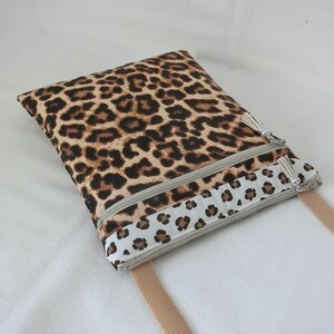 May include: A rectangular crossbody bag with a leopard print pattern in shades of brown and black. The bag features two zippered compartments, a beige strap, and a decorative tassel on each zipper. The bottom section has a white border with a smaller leopard print.