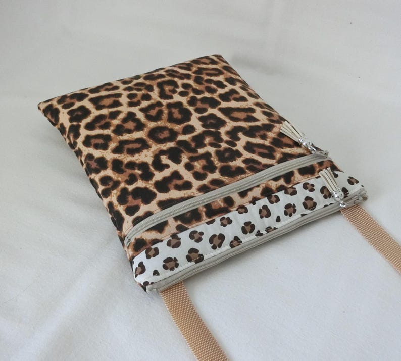 May include: A rectangular crossbody bag with a leopard print pattern in shades of brown and black. The bag features two zippered compartments, a beige strap, and a decorative tassel on each zipper. The bottom section has a white border with a smaller leopard print.