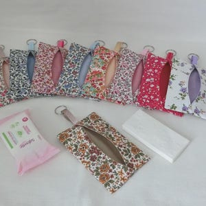 May include: A selection of floral fabric tissue holders in assorted colours, each with a keyring attachment. A packet of wet wipes and a stack of white tissues are also visible. The tissue holders have a slit opening for easy access.