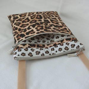 May include: A rectangular crossbody bag with a leopard print pattern. The bag features a beige zipper and a matching beige strap. The interior lining is white with a smaller leopard print. The bag is designed with a tassel zipper pull.