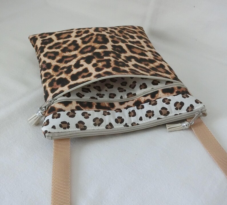 May include: A rectangular crossbody bag with a leopard print pattern. The bag features a beige zipper and a matching beige strap. The interior lining is white with a smaller leopard print. The bag is designed with a tassel zipper pull.
