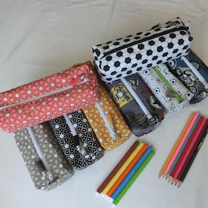 Puede incluir: Una colección de estuches rectangulares de tela con varios patrones, incluyendo formas geométricas, balones de fútbol y gráficos de motocicletas, cada uno con cierre de cremallera y tirador de borla, acompañados de varios lápices de colores.