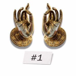 Puede incluir: Dos esculturas decorativas de manos doradas, en un gesto de bendición. Cada mano está montada sobre una base circular con un diseño ornamentado. Una pequeña etiqueta blanca debajo dice "#1".