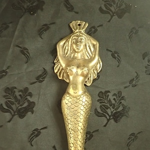 Może przedstawiać: Złota figurka syreny o szczegółowym wzornictwie. Syrena ma długie, falujące włosy, muszelkowy biustonosz i łuskowaty ogon. Figurka stoi na czarnym tle.