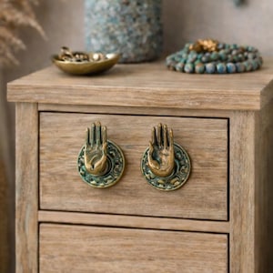 May include: A light-colored wooden nightstand with two drawers, adorned with hand-shaped brass drawer pulls. A lamp with a textured blue shade and base sits on top, along with decorative beads and a small brass dish. The scene evokes a bohemian bedroom aesthetic.