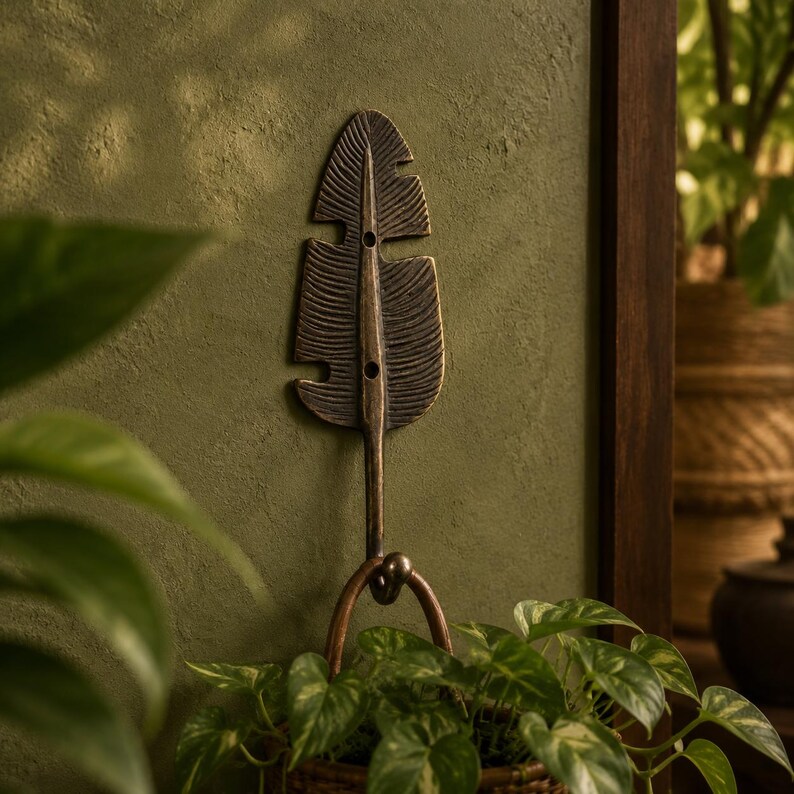 Antique brass monstera leaf wall hook mounted on a dark green wall with a potted plant beside it