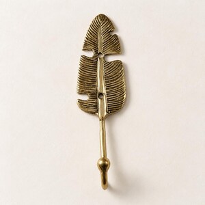 Single solid brass banana leaf hook mounted on a cream colored wall, close view of leaf shape and hook design