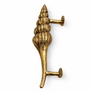 May include: A golden-colored, seashell-shaped door handle. The handle has a textured surface and features a spiral design at the top, with two horizontal extensions for mounting. The overall design is reminiscent of marine life.