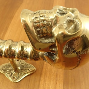 May include: A brass skull-shaped door handle. The handle features a detailed skull design with visible teeth and stitched detailing. The handle is mounted on a decorative base and has a textured, ribbed shaft.