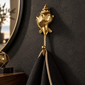 Brass shell spiral hook mounted on a dark wall near a round mirror, holding a black towel