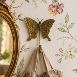 May include: A brass butterfly wall hook with intricate wing details hangs on a floral wallpaper background. A beige tote bag with a floral pattern is suspended from the hook. A gold-framed mirror is visible on the left.