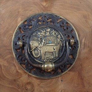 May include: An ornate brass door knocker with an elephant design. The knocker has a detailed elephant in the centre, surrounded by an openwork border. It is mounted on a wooden surface. The knocker's diameter is approximately 15 cm.
