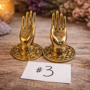 May include: Two gold-colored hand-shaped decorative objects with detailed finger and palm features, mounted on ornate circular bases. A small white card with the number "3" is in front of them. The background includes flowers and a candle.