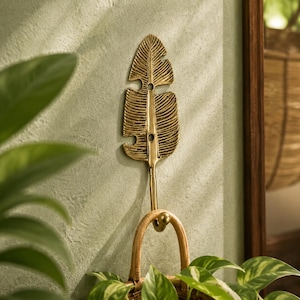 Solid brass banana leaf hook mounted on a textured wall surrounded by trailing green house plants, boho jungalow home decor
