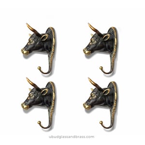 May include: Four bronze-colored bull head wall hooks. Each hook features a detailed bull head with golden horns and accents, mounted on a decorative backplate with a curved hook below. The website address ubudglassandbrass.com is at the bottom.
