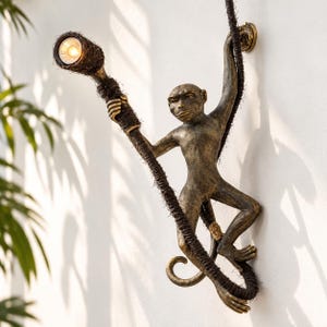May include: A wall-mounted lamp featuring a bronze-toned monkey figure climbing a dark, textured rope. The monkey holds a light fixture at the end of the rope, which emits a warm glow. The design is secured to the wall with a circular mount.
