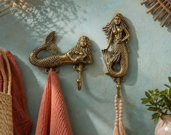 May include: Two antique brass mermaid wall hooks. One mermaid is horizontal, the other vertical. A coral-coloured towel hangs from the horizontal hook. The vertical hook has a beaded tassel. The hooks are mounted on a light blue wall.