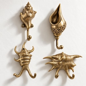 May include: A set of four wall-mounted hooks finished in a metallic gold tone, each shaped like a different seashell. The hooks feature intricate textures and details, with two hooks extending from the base of the two bottom shell designs.