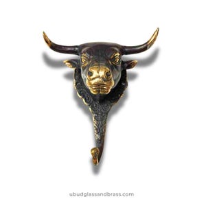 May include: A bronze-colored bull head wall hook with a gold-colored snout and horns. The hook has an ornate, decorative design and is suitable for hanging items. The website address "ubudglassandbrass.com" is visible at the bottom of the image.