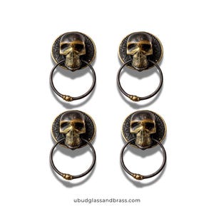 May include: Four antique brass skull door knockers. Each knocker features a skull design with a ring handle. The knockers have a dark brass finish with gold accents. The website address ubudglassandbrass.com is visible at the bottom.