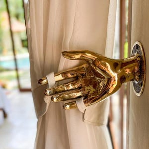 May include: A gold-colored hand-shaped curtain tieback. The hand is attached to a circular wall mount. The curtain is a light beige color and is held in place by white fabric straps around the fingers.