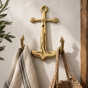 Brass anchor wall hook mounted on a cream wall holding a striped towel and woven bag, nautical maritime coat hook for coastal beach house decor