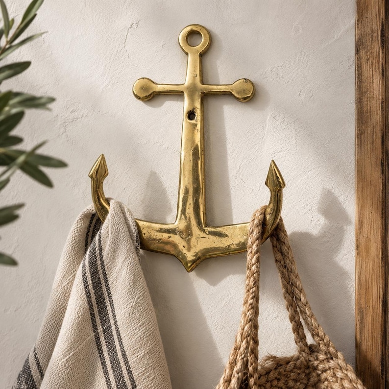 Brass anchor wall hook mounted on a cream wall holding a striped towel and woven bag, nautical maritime coat hook for coastal beach house decor