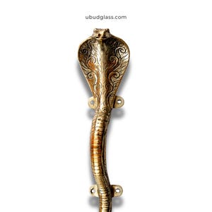 May include: A brass cobra-shaped door handle with detailed engravings on the hood. The handle has a curved design and is mounted with screw holes. The handle is a golden colour and has a textured surface. The website address ubudglass.com is visible.