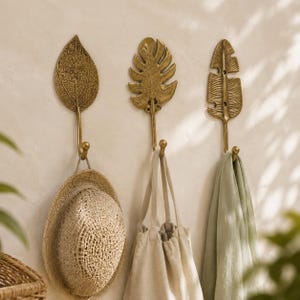 Set of three brass leaf wall hooks in an entryway holding a woven hat, cloth tote bag, and linen towel