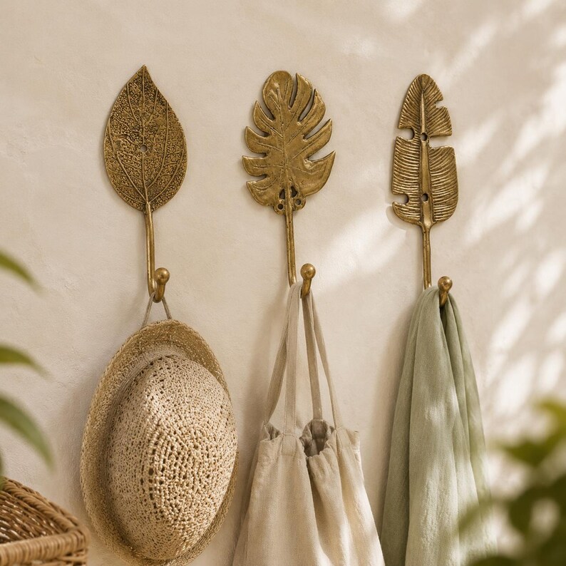 Set of three brass leaf wall hooks in an entryway holding a woven hat, cloth tote bag, and linen towel