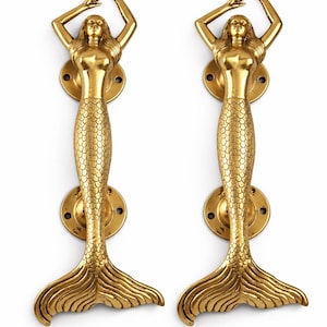 Könnte beinhalten: Zwei goldfarbene Türgriffe in Form einer Meerjungfrau. Jeder Griff zeigt eine Meerjungfrau mit erhobenen Armen, einem schuppenartigen Schwanz und einer kreisförmigen Montageplatte. Die Griffe sind aus Metall.