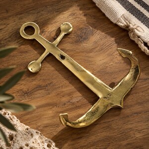 Brass anchor wall hook laid on a wood surface with striped linen and lace fabric nearby, nautical brass hardware coastal decor
