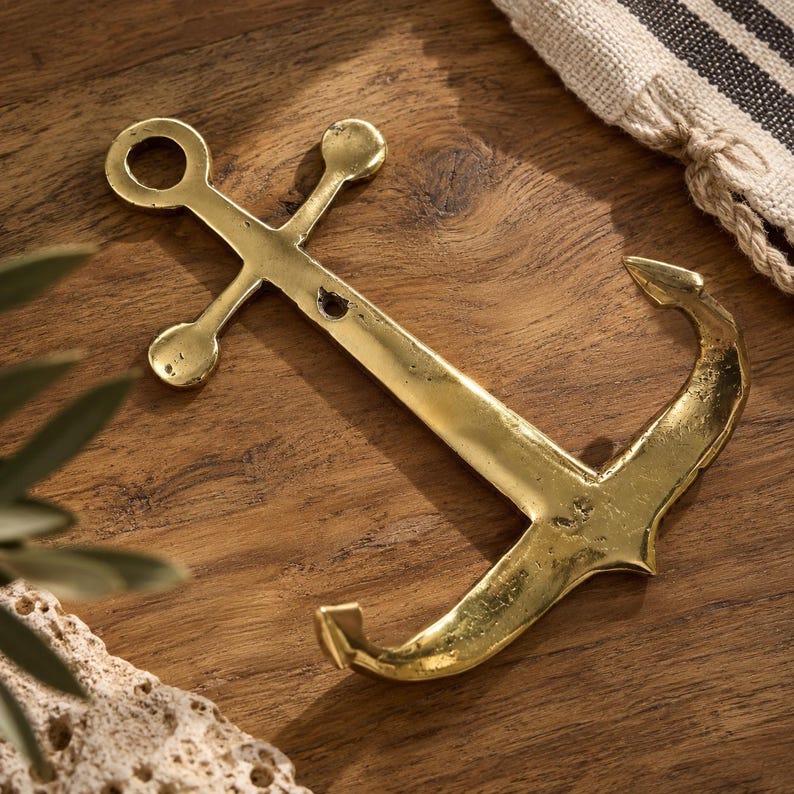 Brass anchor wall hook laid on a wood surface with striped linen and lace fabric nearby, nautical brass hardware coastal decor