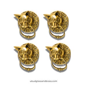 May include: Four gold-colored cat-head door knockers. Each knocker features a detailed cat face with a ring pull. The knockers are mounted on ornate circular backplates. The website address ubudglassandbrass.com is at the bottom.