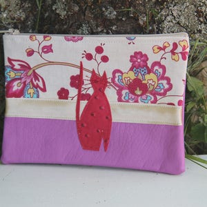 Peut inclure: Une pochette zippée à motif floral blanc et rose avec une applique de chat en feutre rouge. La pochette a une bande de tissu crème entre le motif floral et le tissu rose.