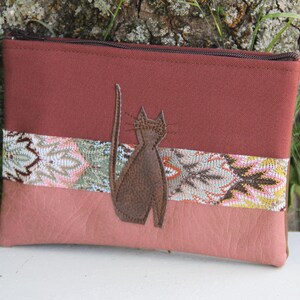 Peut inclure: Une pochette zippée bordeaux avec une applique de chat en cuir marron. La pochette est ornée d'une bande de tissu à motifs rose et blanc.