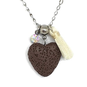 Peut inclure: Un collier avec un pendentif en forme de cœur en roche volcanique brune, une chaîne argentée et un pompon blanc. Le pendentif a un charme de cristal scintillant.