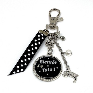 Könnte beinhalten: Ein Schlüsselanhänger aus schwarz-weißem gepunktetem Band mit einem silbernen Anhänger, auf dem "Bientôt Tata!" mit einem Storch und Sternen steht. Der Anhänger ist an einer silbernen Kette mit einem silbernen Blumenanhänger und einer Perle befestigt.