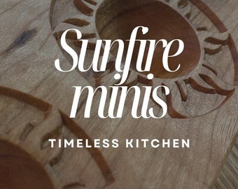 May include: A wooden ravioli mold with three sun-shaped impressions. The mold is labeled "Sunfire minis" and "Timeless Kitchen". The bottom of the mold is labeled "Noce WoodShop".