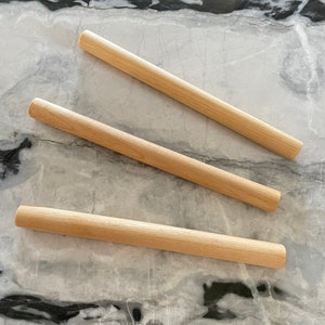 May include: Three wooden rolling pins of varying lengths on a white and black marble surface.
