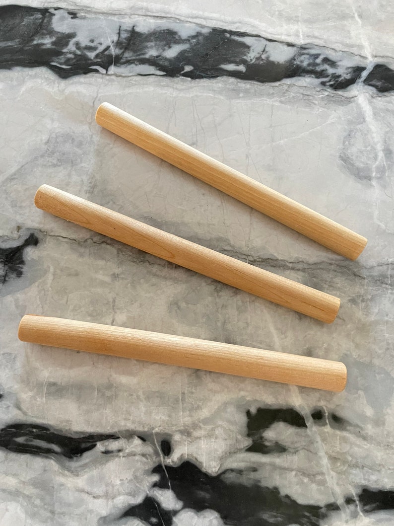 May include: Three wooden rolling pins of varying lengths on a white and black marble surface.