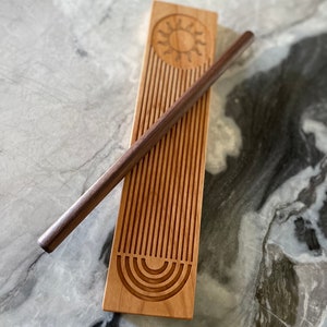 May include: A wooden rolling pin with a sun and rainbow design. The rolling pin is made of light wood and has a dark wood handle. The sun and rainbow design is carved into the wood.